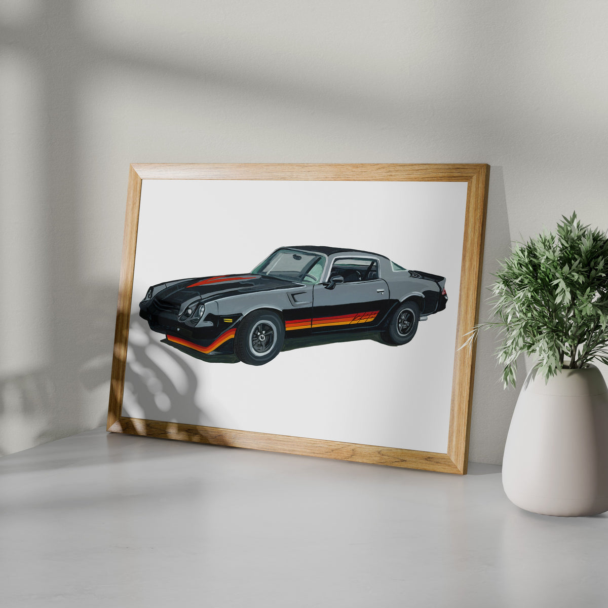 Black Chevrolet Camaro Z28 art print in a wooden frame beside a potted plant