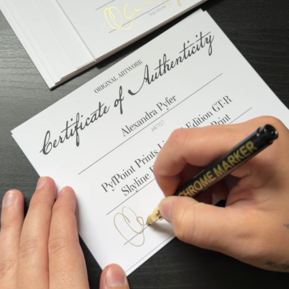Alex hand-signing a Certificate of Authenticity in gold ink for the limited edition Nissan Skyline GT-R R34 art print, with a stack of signed certificates beside her