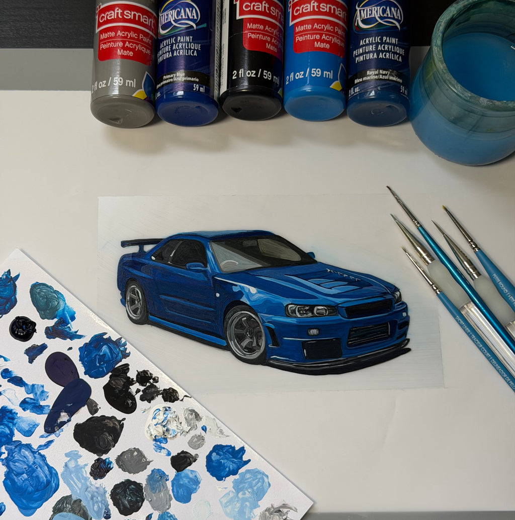 The original Nissan Skyline GT-R R34 acrylic painting in progress on Alex's desk, surrounded by paint bottles, brushes and a mixing palette, before the digital background was added