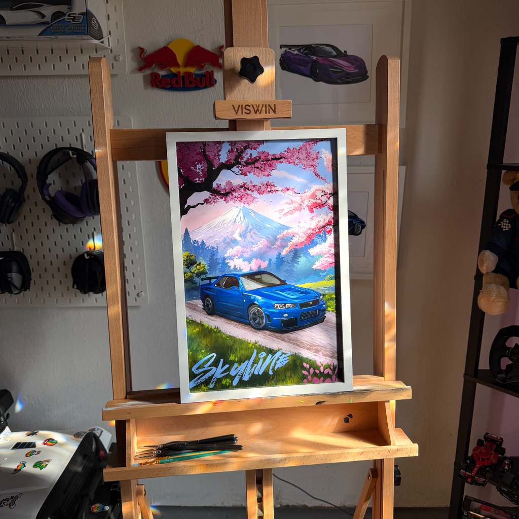 Framed print of a blue Nissan Skyline GT-R R34 on a mountain road under cherry blossoms with Mt. Fuji behind it, displayed on a wooden easel in a garage