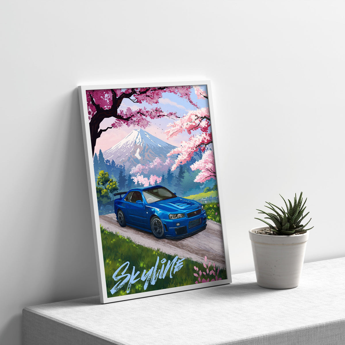 Blue Nissan Skyline GT-R R34 art print in a white frame leaning on a shelf beside a small potted plant