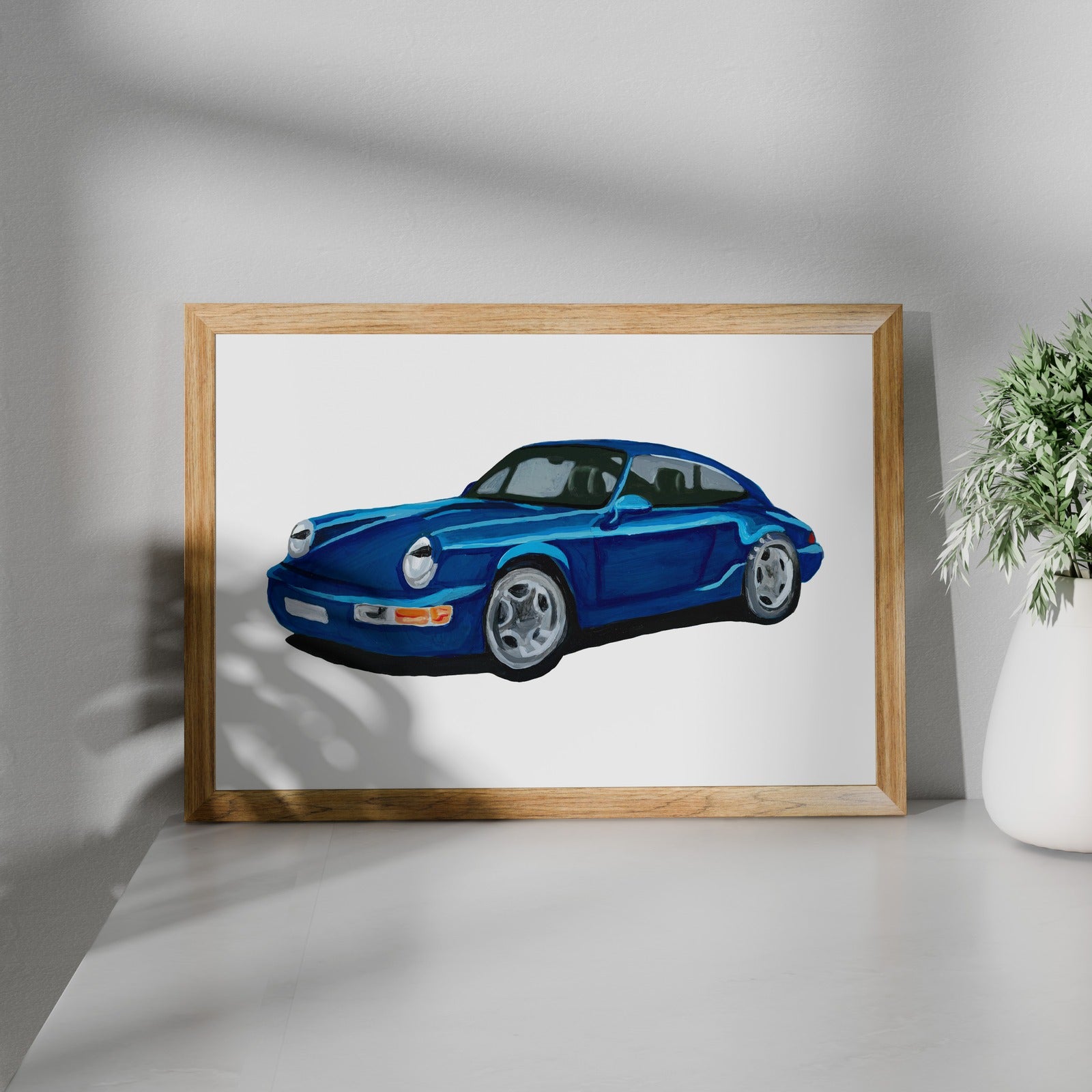 Blue classic sports car art print in a wooden frame beside a potted plant