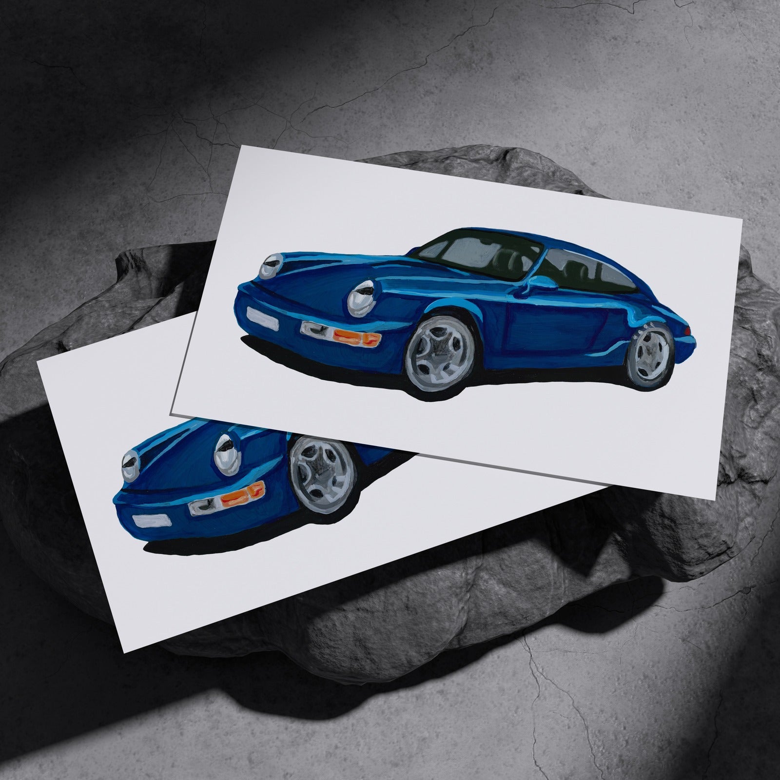 Two blue classic sports car art prints resting on a dark stone surface