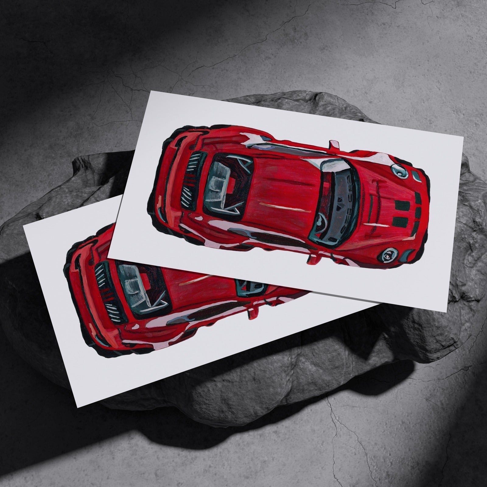Two red sports car art prints resting on a dark stone surface