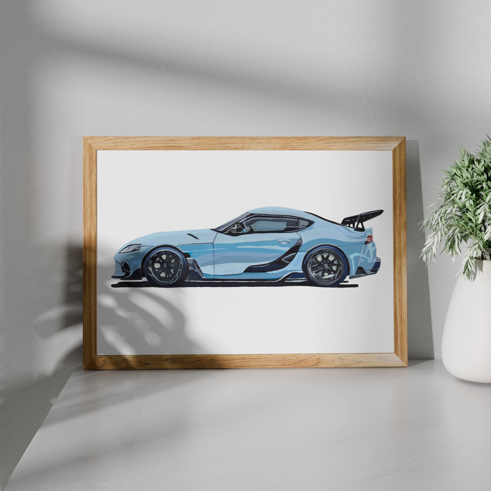 Light blue sports car art print in a wooden frame beside a potted plant